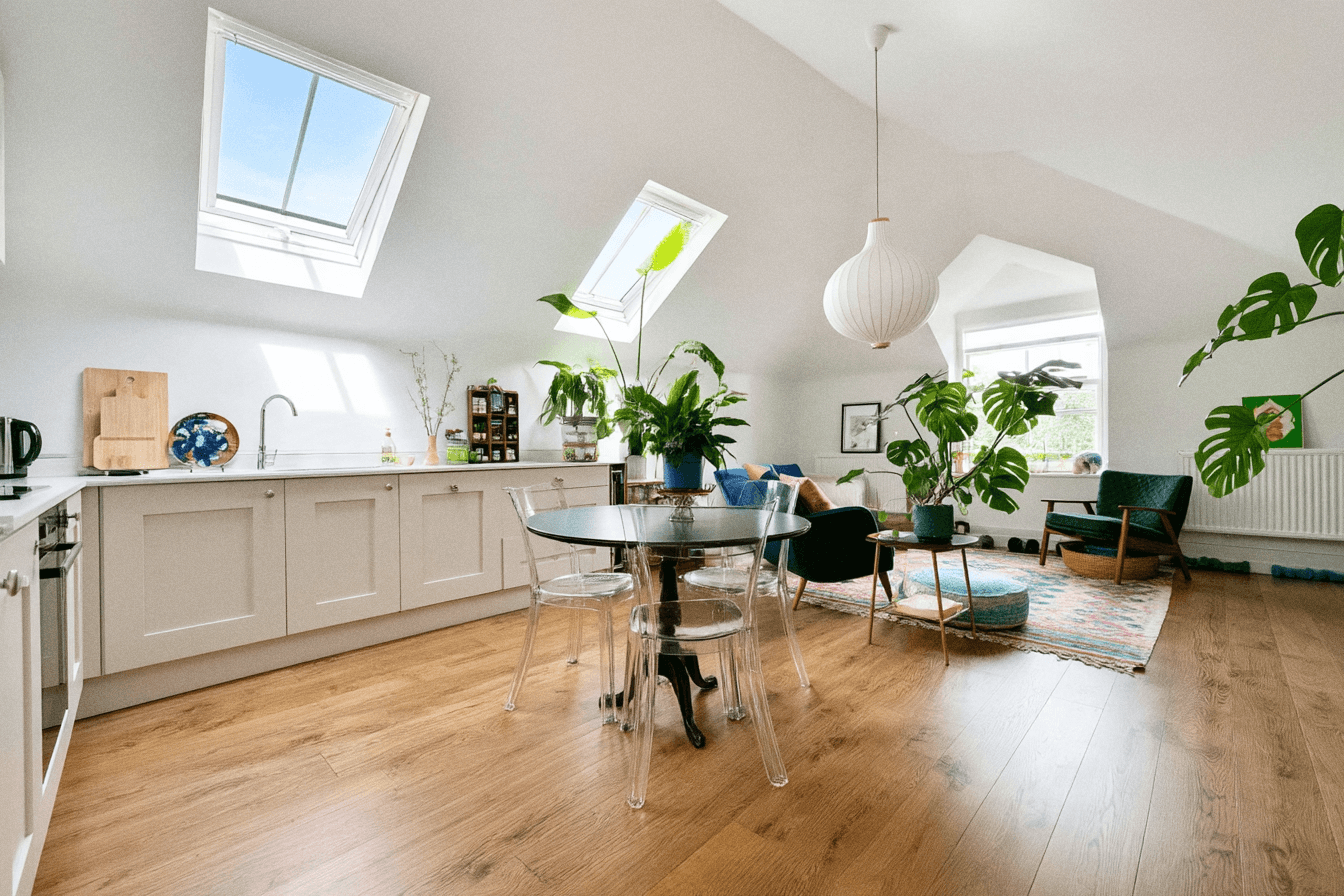 A bright open-plan attic flat with skylights, a shaker-style kitchen, a round dining table with clear chairs and plants throughout
